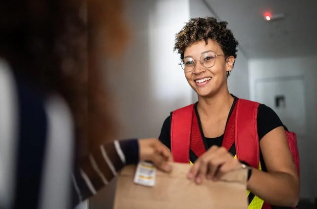 Smiling delivery driver hands a package to a customer, highlighting the evolving landscape of first-party and third-party food delivery services.